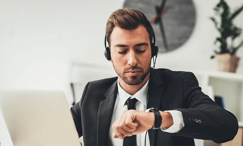 An individual wearing a suit and headset seated at a desk, using a laptop and checking their watch.